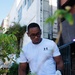 A young man wearing a white Under Armour t-shirt and black-framed glasses is engaged in a community cleanup activity on an urban sidewalk. He is wearing white latex gloves and holding a clear plastic trash bag while using a litter picker tool. The setting appears to be a pedestrian street or alley lined with buildings, plants, and street furniture.
