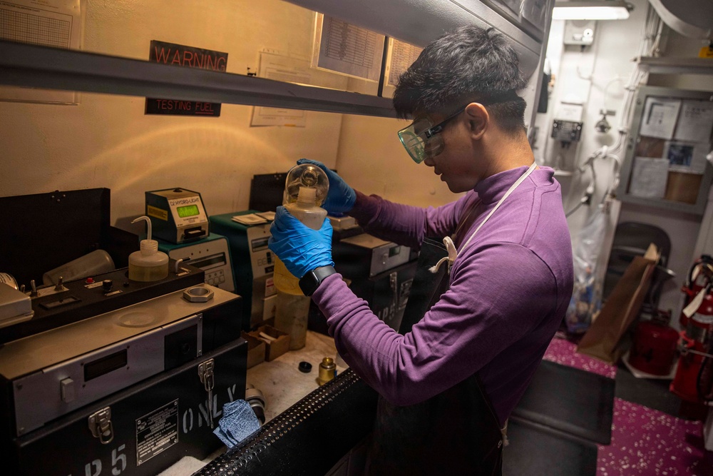 A technician wearing blue nitrile gloves and safety goggles examines a glass flask or bottle containing a liquid sample in what appears to be a fuel testing laboratory. The workspace features multiple testing instruments, warning signs, and safety equipment, with signage indicating 'TESTING FUEL' visible in the background. The setting appears to be aboard a naval vessel based on the confined space and industrial interior design.