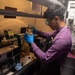 A technician wearing blue nitrile gloves and safety goggles examines a glass flask or bottle containing a liquid sample in what appears to be a fuel testing laboratory. The workspace features multiple testing instruments, warning signs, and safety equipment, with signage indicating 'TESTING FUEL' visible in the background. The setting appears to be aboard a naval vessel based on the confined space and industrial interior design.