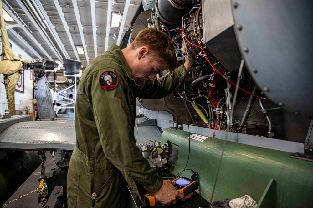A military technician in an olive green flight suit is performing maintenance on an aircraft engine in what appears to be a hangar bay, likely aboard a naval vessel. The mechanic is using a handheld electronic diagnostic device while working on an exposed engine compartment with visible wiring, hoses, and mechanical components. The setting suggests an enclosed aircraft maintenance facility with multiple aircraft visible in the background.