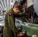 A military technician in an olive green flight suit is performing maintenance on an aircraft engine in what appears to be a hangar bay, likely aboard a naval vessel. The mechanic is using a handheld electronic diagnostic device while working on an exposed engine compartment with visible wiring, hoses, and mechanical components. The setting suggests an enclosed aircraft maintenance facility with multiple aircraft visible in the background.