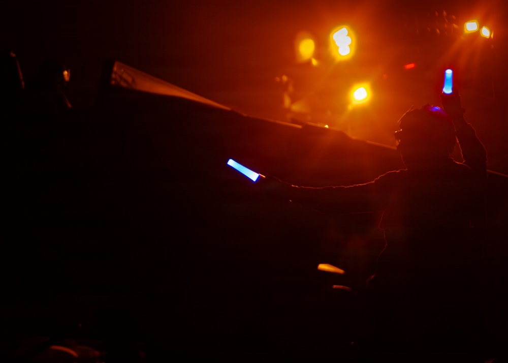 A dark, atmospheric scene showing a silhouetted figure at what appears to be a live concert or nighttime event. The person is holding a blue glow stick and appears to be wearing a blue light accessory on their head. The background is illuminated by warm orange and yellow stage lights with visible haze or fog.