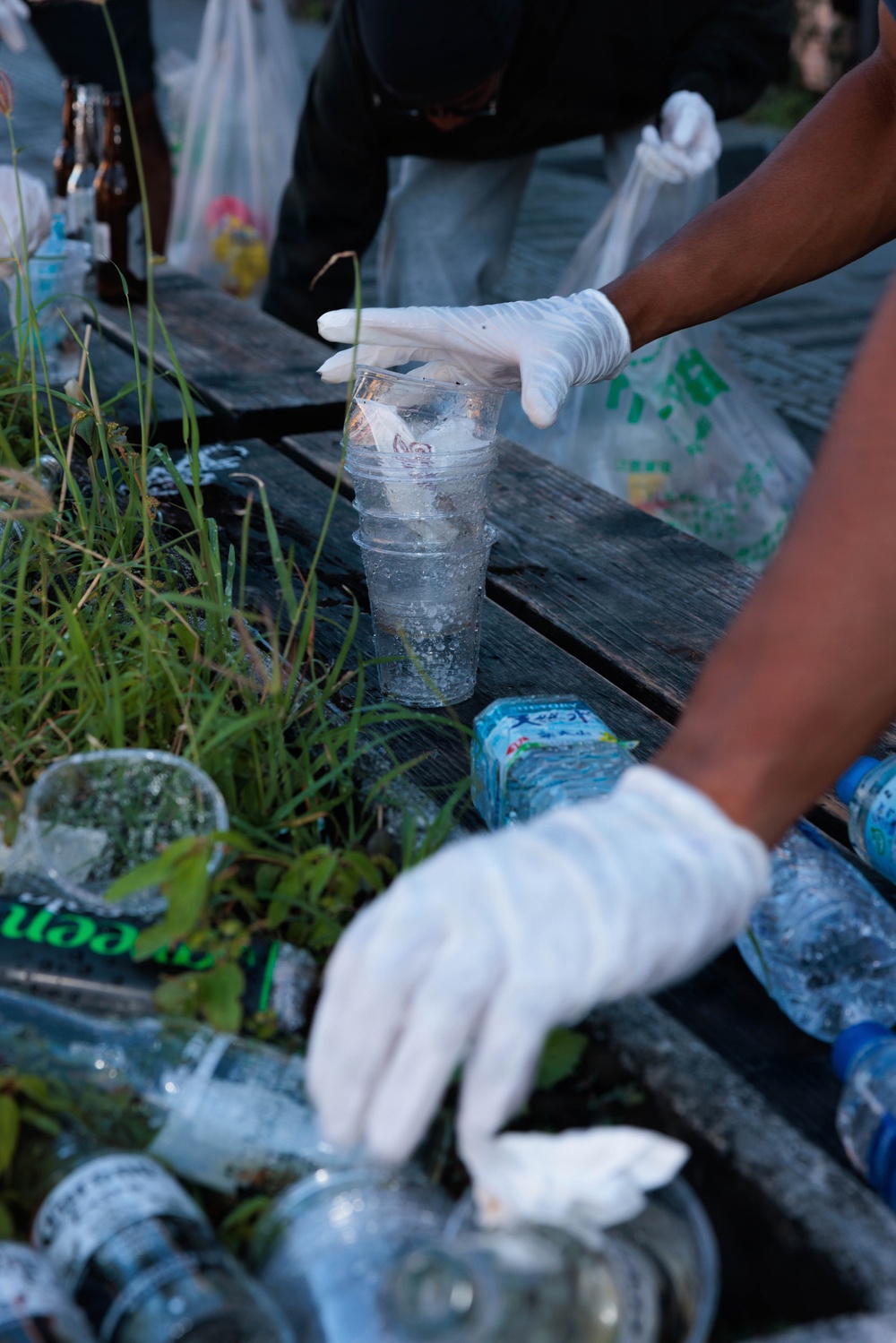 A person wearing white latex gloves is collecting plastic waste and litter from an outdoor surface, likely during a cleanup activity. Multiple plastic cups, bottles, and other debris are visible scattered among grass and vegetation. The scene suggests an organized environmental cleanup effort, possibly in a public outdoor space.