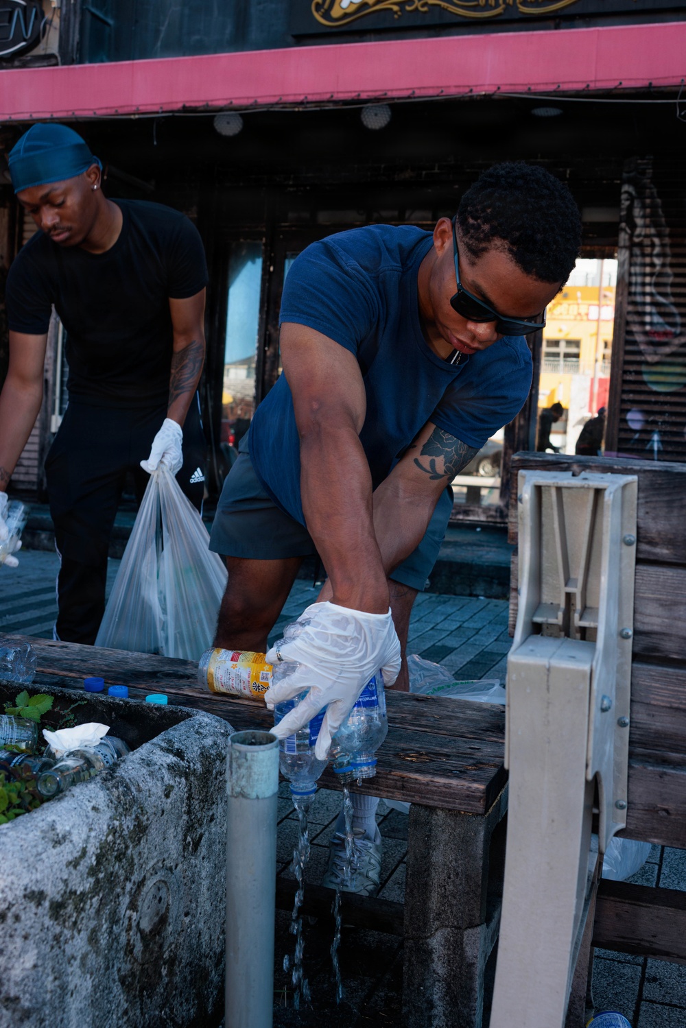 Two men are engaged in a community cleanup effort on an urban street. The foreground figure, wearing a blue t-shirt, dark shorts, and sunglasses, is collecting plastic bottles and debris while wearing white latex gloves. A second man in the background, wearing a black t-shirt and a blue durag, holds a plastic garbage bag with gloved hands.