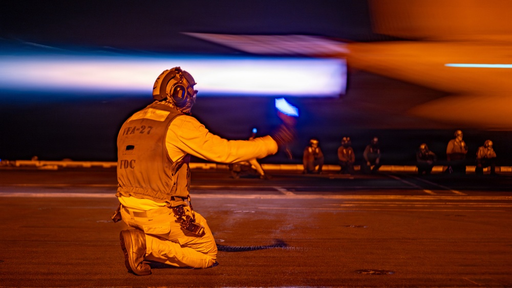 A U.S. Navy flight deck crew member kneels on an aircraft carrier flight deck at night, signaling with a lighted wand as a jet aircraft passes at high speed. The crew member's vest is marked 'VFA-27' and 'FDC' (Flight Deck Coordinator), indicating association with Strike Fighter Squadron 27. Additional deck crew members are visible crouching in the background, illuminated by the warm orange glow of the aircraft's exhaust.