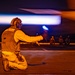 A U.S. Navy flight deck crew member kneels on an aircraft carrier flight deck at night, signaling with a lighted wand as a jet aircraft passes at high speed. The crew member's vest is marked 'VFA-27' and 'FDC' (Flight Deck Coordinator), indicating association with Strike Fighter Squadron 27. Additional deck crew members are visible crouching in the background, illuminated by the warm orange glow of the aircraft's exhaust.