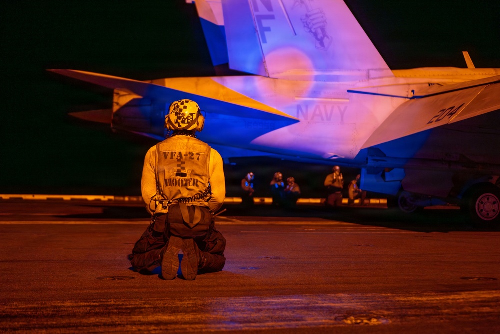 A U.S. Navy flight deck crew member from VFA-27 squadron kneels on a tarmac at night in front of a Navy fighter aircraft, likely an F/A-18 Hornet or Super Hornet bearing the number '204'. The scene is illuminated by warm orange/amber lighting, creating a dramatic nighttime operational atmosphere. Additional crew members are visible in the background near the aircraft.