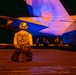 A U.S. Navy flight deck crew member from VFA-27 squadron kneels on a tarmac at night in front of a Navy fighter aircraft, likely an F/A-18 Hornet or Super Hornet bearing the number '204'. The scene is illuminated by warm orange/amber lighting, creating a dramatic nighttime operational atmosphere. Additional crew members are visible in the background near the aircraft.