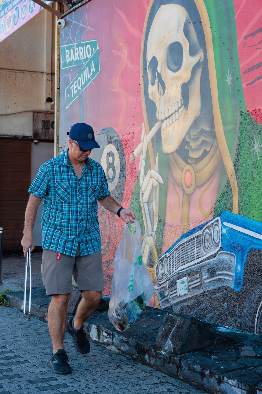A middle-aged man wearing a blue plaid shirt, khaki shorts, and a navy baseball cap walks along a brick sidewalk carrying a plastic bag and a cane or walking stick. He is passing in front of a large, colorful street mural depicting a Santa Muerte (skeletal religious figure) alongside street signs reading 'Barrio' and 'Tequila,' an 8-ball, and a classic lowrider car. The setting appears to be an urban neighborhood, likely in a Latino cultural district.