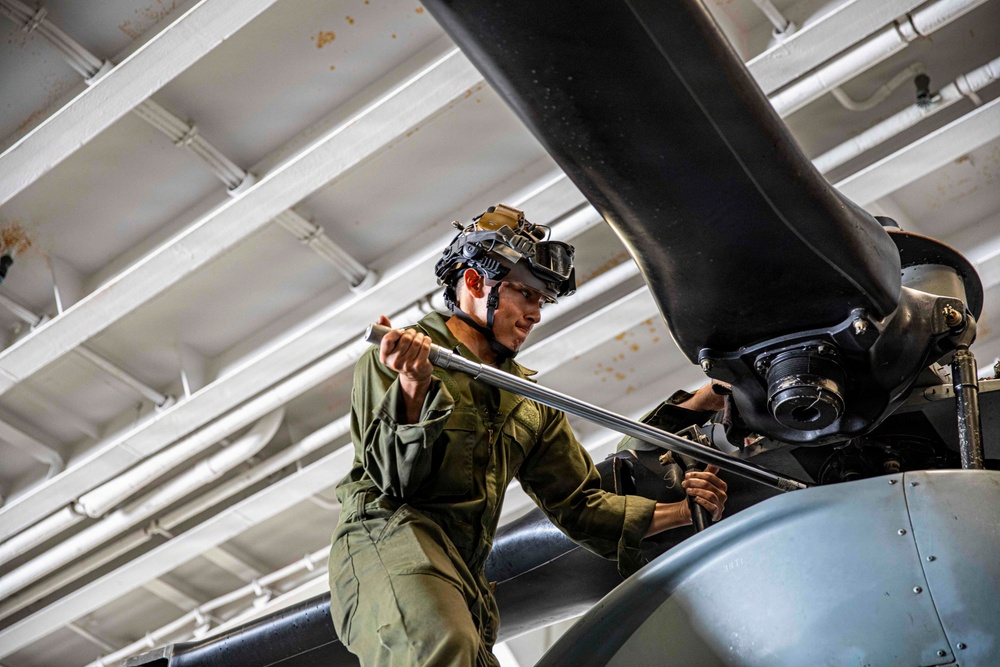 A military technician in olive green flight suit is performing maintenance on a helicopter rotor head assembly, likely inside a ship's hangar bay. The mechanic is using a torque wrench or breaker bar to work on the rotor hub components. The setting appears to be an enclosed naval vessel hangar based on the white-painted structural beams and piping visible overhead.