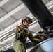 A military technician in olive green flight suit is performing maintenance on a helicopter rotor head assembly, likely inside a ship's hangar bay. The mechanic is using a torque wrench or breaker bar to work on the rotor hub components. The setting appears to be an enclosed naval vessel hangar based on the white-painted structural beams and piping visible overhead.