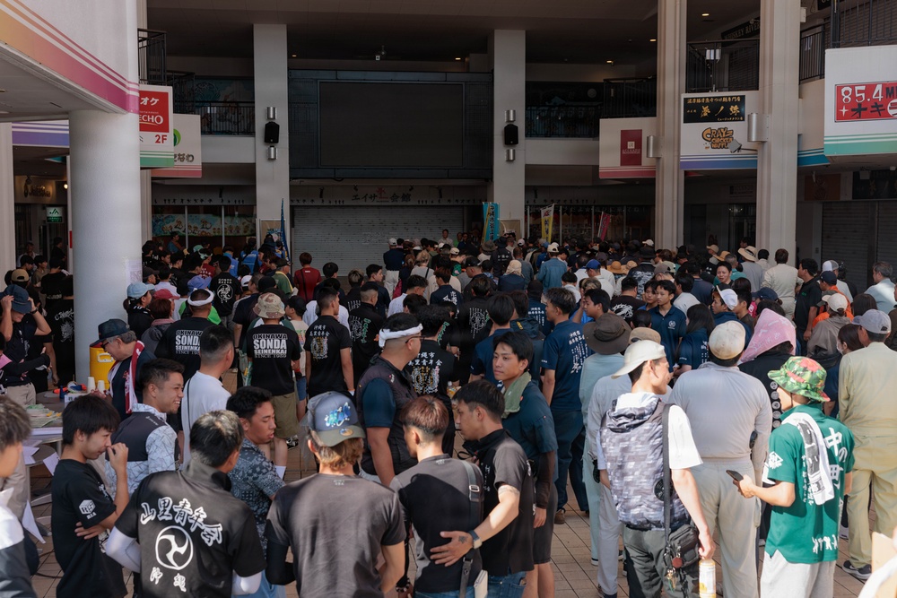 A large crowd of people has gathered in what appears to be a covered outdoor shopping arcade or plaza in Japan. Many attendees are wearing matching black t-shirts with Japanese and English text, suggesting affiliation with various organizations or groups. The gathering appears to be an organized community or cultural event, possibly a festival, rally, or group assembly.