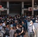 A large crowd of people has gathered in what appears to be a covered outdoor shopping arcade or plaza in Japan. Many attendees are wearing matching black t-shirts with Japanese and English text, suggesting affiliation with various organizations or groups. The gathering appears to be an organized community or cultural event, possibly a festival, rally, or group assembly.