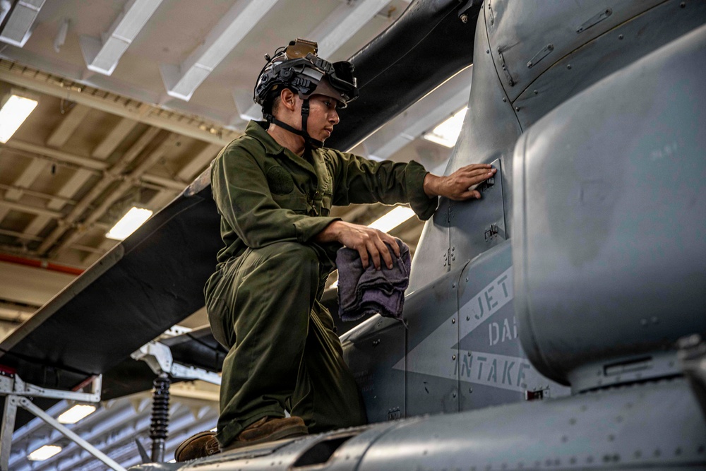 A military aviation technician in olive green flight suit and helmet with night vision goggle mount is performing maintenance on a military aircraft in an indoor hangar bay. The technician is crouched on the aircraft's wing or fuselage, using a cloth to inspect or clean a panel near a jet intake area. The setting appears to be a naval vessel hangar deck based on the overhead lighting and structural elements visible.