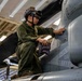 A military aviation technician in olive green flight suit and helmet with night vision goggle mount is performing maintenance on a military aircraft in an indoor hangar bay. The technician is crouched on the aircraft's wing or fuselage, using a cloth to inspect or clean a panel near a jet intake area. The setting appears to be a naval vessel hangar deck based on the overhead lighting and structural elements visible.