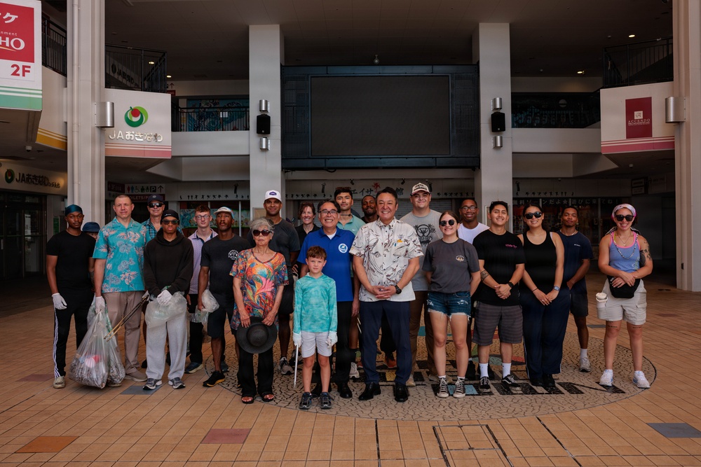 A group of approximately 20 people pose for a group photo in what appears to be a covered outdoor shopping arcade or public plaza in Okinawa, Japan, as indicated by Japanese signage including 'JAおきなわ' (JA Okinawa). Several participants are holding trash bags and litter-picking tools, suggesting this is a community cleanup event. The group appears to include both Japanese locals and what appear to be American military personnel or civilians, indicating a community outreach or volunteer activity.