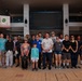 A group of approximately 20 people pose for a group photo in what appears to be a covered outdoor shopping arcade or public plaza in Okinawa, Japan, as indicated by Japanese signage including 'JAおきなわ' (JA Okinawa). Several participants are holding trash bags and litter-picking tools, suggesting this is a community cleanup event. The group appears to include both Japanese locals and what appear to be American military personnel or civilians, indicating a community outreach or volunteer activity.