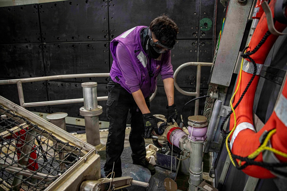 A worker in a purple vest and black cargo pants is performing maintenance or mechanical work on piping or valve equipment aboard what appears to be a vessel or industrial platform. The individual is wearing safety goggles, a face mask, and black gloves while bending over to work on a purple/lavender-colored pipe fitting or valve assembly. Orange life-saving equipment is visible on the right side, confirming a maritime environment.