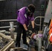 A worker in a purple vest and black cargo pants is performing maintenance or mechanical work on piping or valve equipment aboard what appears to be a vessel or industrial platform. The individual is wearing safety goggles, a face mask, and black gloves while bending over to work on a purple/lavender-colored pipe fitting or valve assembly. Orange life-saving equipment is visible on the right side, confirming a maritime environment.