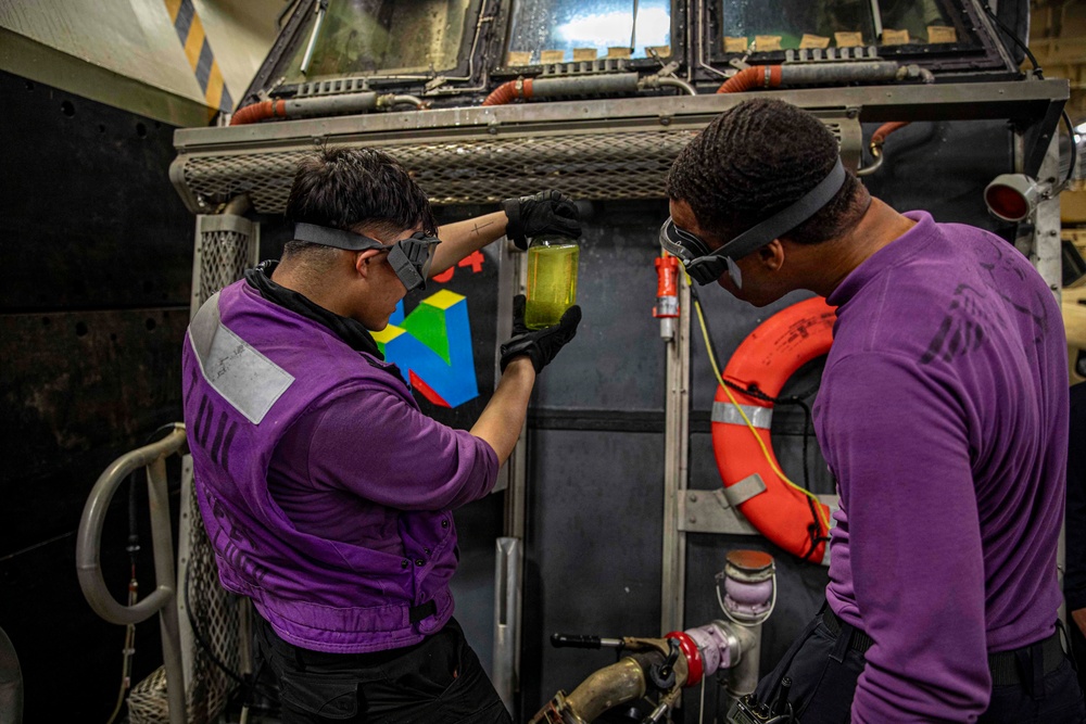 Two personnel wearing purple jerseys and safety goggles are examining a clear container filled with yellow-green liquid, likely conducting a fuel quality check. They are working in what appears to be a naval vessel's hangar or deck area, surrounded by industrial equipment. The purple jerseys indicate they are aviation fuels personnel (known as 'grapes') aboard a U.S. Navy aircraft carrier.