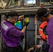 Two personnel wearing purple jerseys and safety goggles are examining a clear container filled with yellow-green liquid, likely conducting a fuel quality check. They are working in what appears to be a naval vessel's hangar or deck area, surrounded by industrial equipment. The purple jerseys indicate they are aviation fuels personnel (known as 'grapes') aboard a U.S. Navy aircraft carrier.