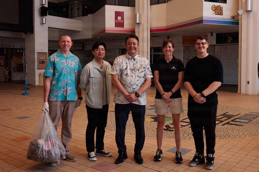 Five individuals are posing for a group photo inside what appears to be a shopping mall or commercial center. The group consists of three men and two women standing together in a semi-formal arrangement, with one person holding a clear plastic bag containing collected items. The setting suggests a community or volunteer event, possibly a cleanup activity.