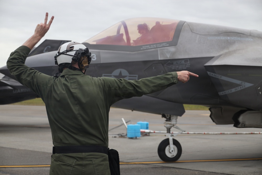 A military flight deck crew member in a green flight suit and white helmet is directing an F-35 fighter jet on a tarmac. The marshal is using hand signals — raising two fingers with one hand and pointing with the other — to guide the aircraft. The jet's cockpit is open with a pilot visible inside, and the aircraft bears 'MARINES' markings.