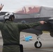 A military flight deck crew member in a green flight suit and white helmet is directing an F-35 fighter jet on a tarmac. The marshal is using hand signals — raising two fingers with one hand and pointing with the other — to guide the aircraft. The jet's cockpit is open with a pilot visible inside, and the aircraft bears 'MARINES' markings.