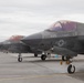 Multiple F-35 stealth fighter jets are lined up on a military airfield tarmac. The aircraft display U.S. military markings including the star-and-bar insignia, with visible tail numbers 225 and 133. Personnel can be seen in the background near the aircraft line.