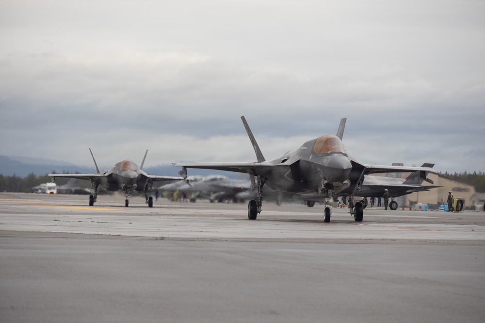 Multiple F-35 Lightning II stealth fighter jets are taxiing on an airfield tarmac in what appears to be a cold, overcast environment. At least two aircraft are clearly visible in the foreground with additional aircraft visible in the background. Ground crew personnel and support vehicles are visible along the flight line near hangar structures.