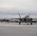 Multiple F-35 Lightning II stealth fighter jets are taxiing on an airfield tarmac in what appears to be a cold, overcast environment. At least two aircraft are clearly visible in the foreground with additional aircraft visible in the background. Ground crew personnel and support vehicles are visible along the flight line near hangar structures.