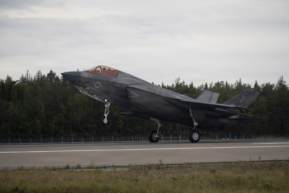 A U.S. Marine Corps F-35B Lightning II stealth fighter jet is captured in the moment of takeoff or landing on a runway, with landing gear fully extended. The aircraft bears visible U.S. military markings including a star-and-bar insignia and tail code 'CE' with number '113'. The setting appears to be a northern or boreal environment based on the dense conifer tree line in the background under overcast skies.