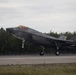 A U.S. Marine Corps F-35B Lightning II stealth fighter jet is captured in the moment of takeoff or landing on a runway, with landing gear fully extended. The aircraft bears visible U.S. military markings including a star-and-bar insignia and tail code 'CE' with number '113'. The setting appears to be a northern or boreal environment based on the dense conifer tree line in the background under overcast skies.