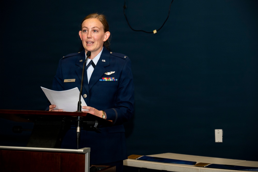 A female U.S. Air Force officer in dress blues is speaking at a podium while holding papers. She is wearing a pilot badge and several service ribbons on her uniform. The setting appears to be an indoor military ceremony or formal briefing.