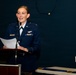 A female U.S. Air Force officer in dress blues is speaking at a podium while holding papers. She is wearing a pilot badge and several service ribbons on her uniform. The setting appears to be an indoor military ceremony or formal briefing.