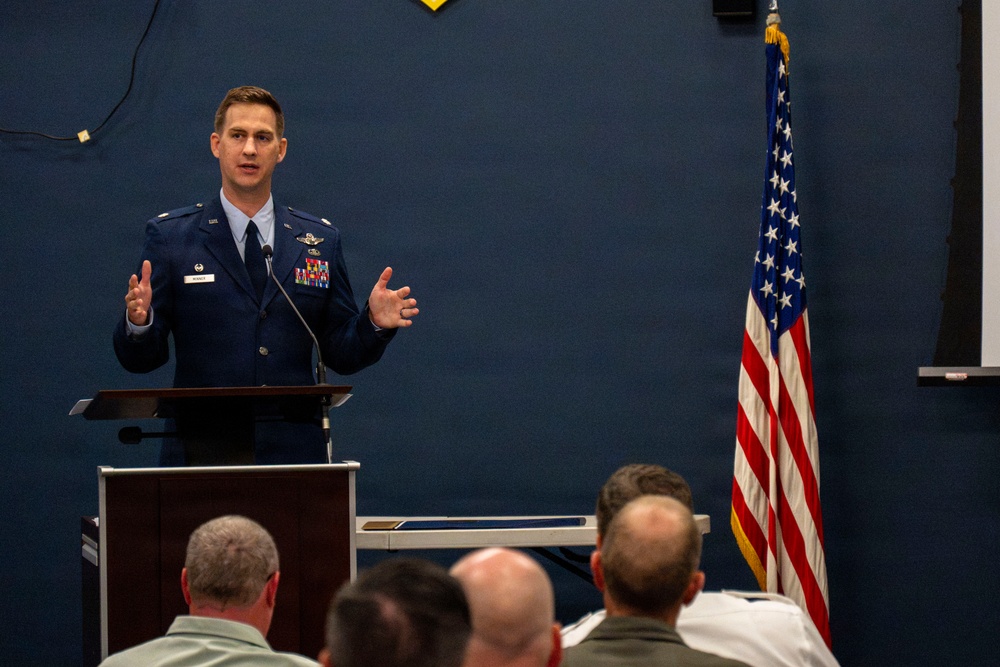 A U.S. Air Force officer in dress blues is delivering a speech or briefing at a podium to an audience of seated military personnel. The setting appears to be an official military ceremony or briefing room, indicated by the American flag and blue-painted walls. The speaker is gesturing with both hands, suggesting an engaged and active presentation.
