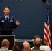 A U.S. Air Force officer in dress blues is delivering a speech or briefing at a podium to an audience of seated military personnel. The setting appears to be an official military ceremony or briefing room, indicated by the American flag and blue-painted walls. The speaker is gesturing with both hands, suggesting an engaged and active presentation.