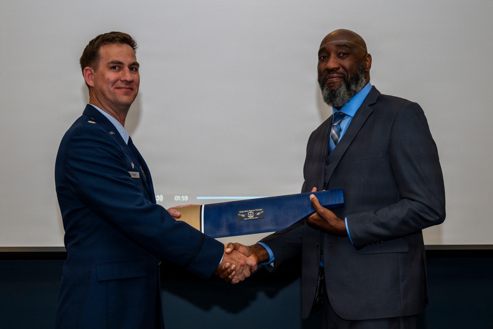 Two men are shaking hands during what appears to be a formal award or recognition ceremony. The man on the left is wearing a U.S. Air Force dress uniform, while the man on the right is wearing a dark suit and blue shirt. They are jointly holding a blue certificate or award folder between them.