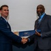 Two men are shaking hands during what appears to be a formal award or recognition ceremony. The man on the left is wearing a U.S. Air Force dress uniform, while the man on the right is wearing a dark suit and blue shirt. They are jointly holding a blue certificate or award folder between them.