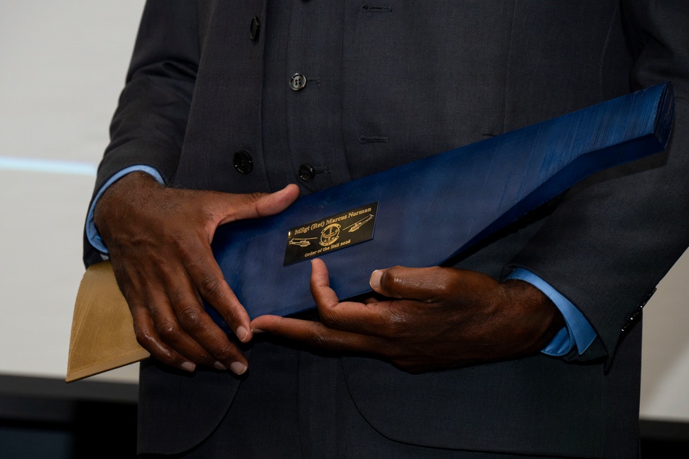 A person wearing a dark charcoal suit with a light blue dress shirt is holding a blue presentation folder or award portfolio. A black and gold nameplate on the folder reads 'MSgt (Ret) Marcus Norman' and 'Order of the Nail 2025'. The image is tightly cropped to focus on the hands and the item being held.