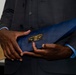 A person wearing a dark charcoal suit with a light blue dress shirt is holding a blue presentation folder or award portfolio. A black and gold nameplate on the folder reads 'MSgt (Ret) Marcus Norman' and 'Order of the Nail 2025'. The image is tightly cropped to focus on the hands and the item being held.