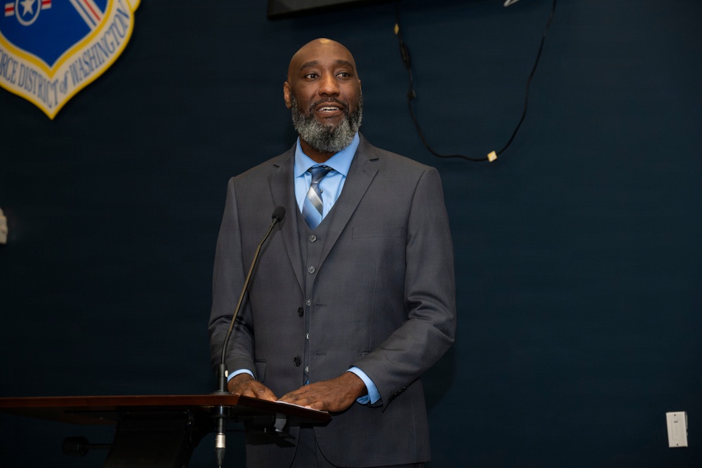 A man in a three-piece gray suit is speaking at a podium with a microphone in front of a dark blue wall. Behind him is a shield-shaped emblem reading 'Air Force District of Washington.' The setting appears to be a formal official or military-affiliated event.