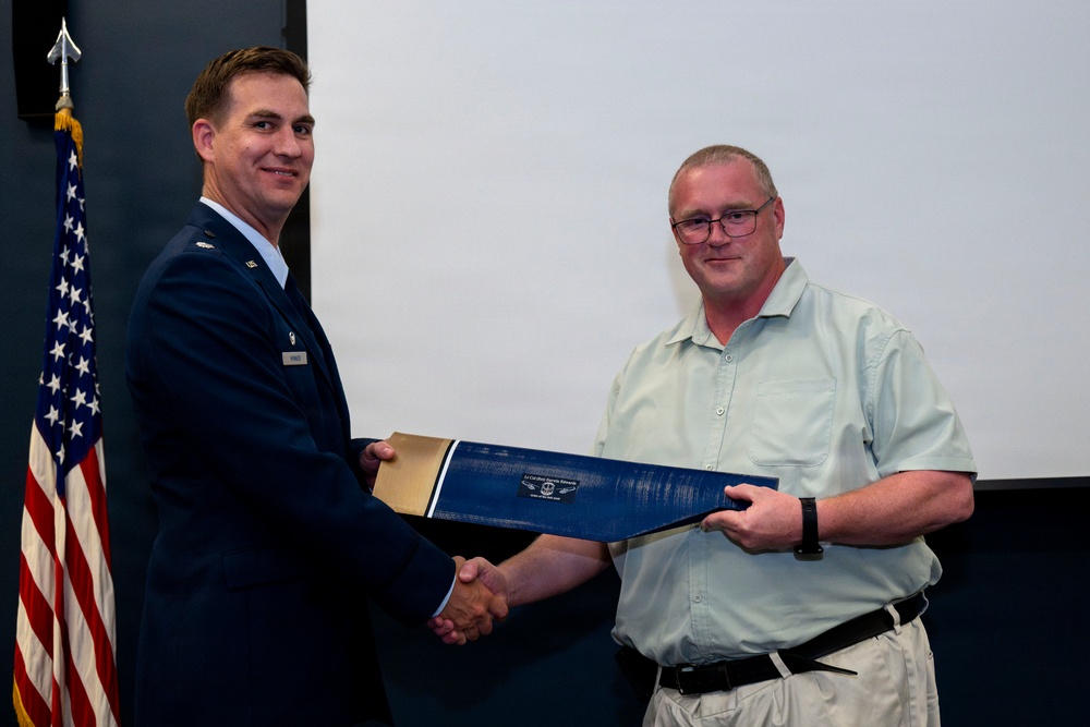 A U.S. Air Force officer in dress uniform is presenting a certificate or award folder to a civilian man during a formal ceremony. Both individuals are shaking hands while jointly holding a blue presentation folder. An American flag is visible in the background to the left.