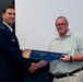 A U.S. Air Force officer in dress uniform is presenting a certificate or award folder to a civilian man during a formal ceremony. Both individuals are shaking hands while jointly holding a blue presentation folder. An American flag is visible in the background to the left.