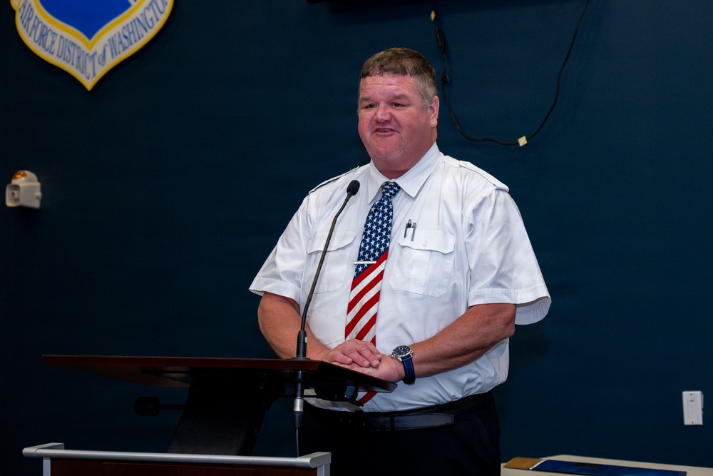 A man in a white short-sleeve uniform shirt and American flag necktie stands at a wooden podium with a microphone, appearing to speak or present. Behind him is a dark blue wall displaying the Air Force District of Washington shield/emblem. The setting appears to be an official military or government briefing room.