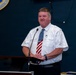 A man in a white short-sleeve uniform shirt and American flag necktie stands at a wooden podium with a microphone, appearing to speak or present. Behind him is a dark blue wall displaying the Air Force District of Washington shield/emblem. The setting appears to be an official military or government briefing room.