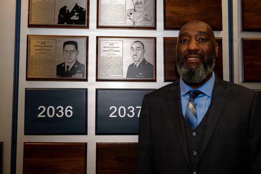 A man in a dark suit with a blue shirt and striped tie stands in front of a military hall of fame or recognition wall. The wall displays framed plaques with portraits and biographical information of military honorees, with year markers including 2026, 2036, and 2037. One visible plaque identifies 'Master Sergeant (Retired) Marcus A. Norman' with a year of induction of 2026.