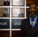 A man in a dark suit with a blue shirt and striped tie stands in front of a military hall of fame or recognition wall. The wall displays framed plaques with portraits and biographical information of military honorees, with year markers including 2026, 2036, and 2037. One visible plaque identifies 'Master Sergeant (Retired) Marcus A. Norman' with a year of induction of 2026.