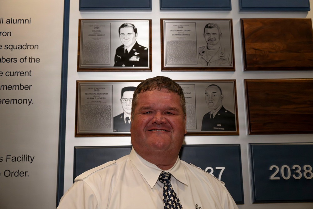A man in a white dress shirt and patterned tie stands smiling in front of a military hall of fame or recognition wall. The wall displays several engraved memorial plaques featuring portrait illustrations and biographical text of military personnel. Numbers '2037' and '2038' are visible on blue plaques in the lower portion of the wall.