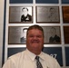 A man in a white dress shirt and patterned tie stands smiling in front of a military hall of fame or recognition wall. The wall displays several engraved memorial plaques featuring portrait illustrations and biographical text of military personnel. Numbers '2037' and '2038' are visible on blue plaques in the lower portion of the wall.